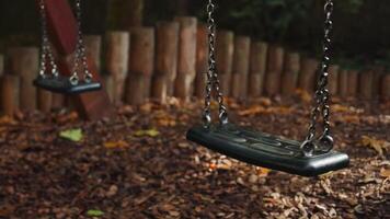Empty black swing on children playground in the park in autumn season. Missing child. Lonely swing in sunny autumn day. Golden foliage in late fall. Beautiful nature. video