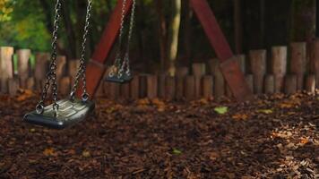 Empty black swing on children playground in the park in autumn season. Missing child. Lonely swing in sunny autumn day. Golden foliage in late fall. Beautiful nature. video
