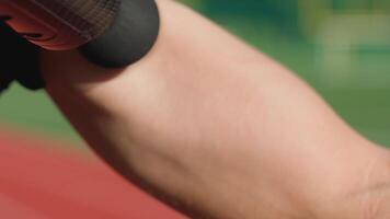 Close-up of male athlete using handheld massage gun on arm muscles for recovery after training session on stadium track. Concept of muscle relaxation and sports recovery video