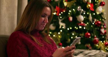 Woman using smartphone with decorated Christmas tree in the background video