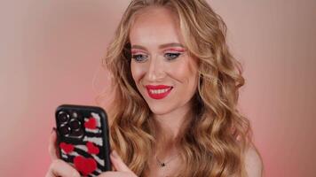 A young woman in a red dress, lipstick takes a selfie with long blond hair and makeup. The girl is smiling and looking at the camera close-up video
