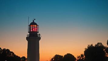 Large, beautiful white lighthouse, with a red-glowing and spinning lens, stands against the backdrop of a beautiful sunset sky. video
