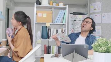 Two Asian workers are unhappy working with hot temperatures at small business office in summer season, uncomfortable with no air conditioner, using fans blowing wind, sitting at desks, and sweating. video