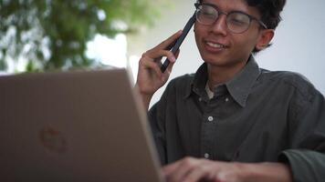 Young Asian man working on his laptop while talking on mobile phone. Business man having phone call while working outdoor video