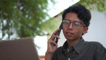 Young Asian man working on his laptop while talking on mobile phone. Business man having phone call while working outdoor video