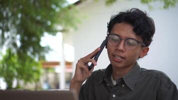 Young Asian man working on his laptop while talking on mobile phone. Business man having phone call while working outdoor video