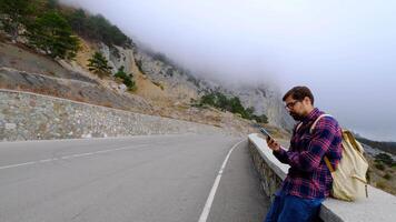 A man is sitting on the road of a mountain and texting a message on his smartphone. He has a large backpack on his shoulders. Hiking in the mountains. video