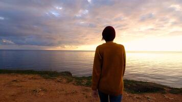 Woman wearing a yellow sweater and a red hat and enjoying beautiful colorful sunset above sea from cape. concepts of the winner, freedom, happiness. hiker exploring nature video