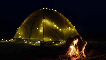 Tent with a garland and burning camp fire at dusk on the beach. Summer adventures and exploration. Selective focus on fire. summer rest , Tent at the background. video