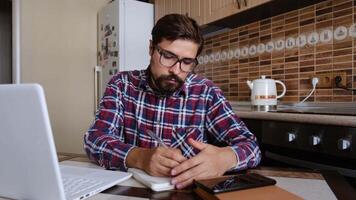 Smiling hipster male student wear glasses elearning study with online tutor teacher , learn language make notes sit at home office desk look at laptop computer video