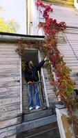 A woman stands on a small balcony, reaching up to touch vibrant red autumn vines growing along the wooden facade of a historical building. The angle emphasizes the height of the structure and the video
