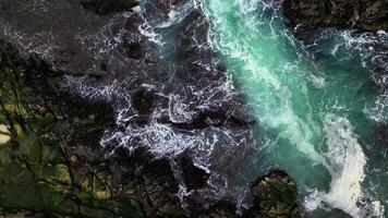 Aerial view the ocean waves crash against the black rocks, creating white foam that contrasts with the dark green seawater. The water flow appears strong, breaking through the rough rock crevices. video