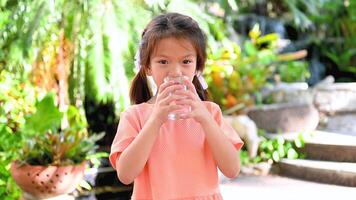 Child Girl drinks clean, cool water from a clear glass with freshness. The background is a garden with a small waterfall and swaying trees. video
