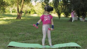 Athletic child girl in VR headset helmet making fitness workout exercises with dumbbells in park video