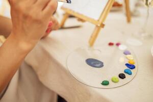 Close-up of woman hands and a palette with paints. The artist holds a brush and mixes acrylic paints. A woman paints on canvas at an easel at a table in the workshop. photo