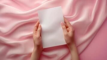 Hands holding blank card invitation mockup against flowing pink satin fabric. Softness, elegance, delicate personal message template advertising image. Mock up product photorealistic photo