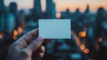 Hand holding blank card mockup against blurred urban skyline at twilight. Soft city lights. Branding messaging businesscard template advertising image. Mock up product photorealistic photo