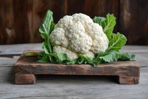 Fresh cauliflower head displayed on wooden cutting board with leafy greens in rustic setting photo