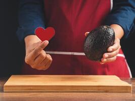 Hand holding a red heart shape symbol with fresh avocado while standing in the kitchen photo