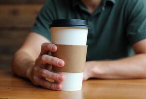 Close-up of a man holding a takeaway coffee cup with a brown sleeve, sitting at a wooden table. photo