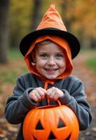 A young boy wearing a witch hat, smiling while holding a pumpkin bucket photo