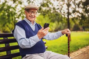 Portrait of happy senior man in park. He is sitting on bench and using smartphone. photo