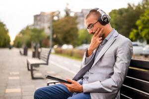 Portrait of thoughtful businessman who is sitting on bench in the city and using digital tablet. photo