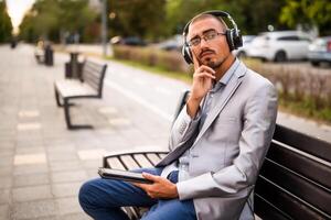 Portrait of thoughtful businessman who is sitting on bench in the city and using digital tablet. photo