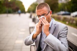 Portrait of businessman who is sitting on bench in the city and blowing nose. He has allergy problem. photo