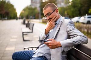 Portrait of pensive businessman who is messaging on phone. Man sitting on bench in the city and using smartphone. photo