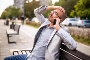 Portrait of businessman in disbelief who is talking on phone. Man sitting on bench in the city and talking on phone. photo