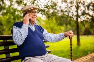 Portrait of happy senior man in park. He is sitting on bench and talking on phone. photo