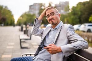 Portrait of businessman in disbelief who is messaging on phone. Man sitting on bench in the city and using smartphone. photo