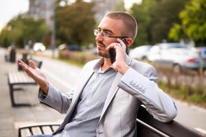 Portrait of businessman in disbelief who is talking on phone. Man sitting on bench in the city and talking on phone. photo