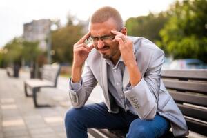 Portrait of overworked and depressed man. He is sitting on bench in the city and having headache. photo