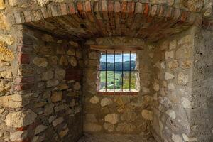 A barred window in a stone wall frames a peaceful view of green hills, fields, and distant forests photo