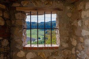 A barred window in a stone wall frames a peaceful view of green hills, fields, and distant forests photo