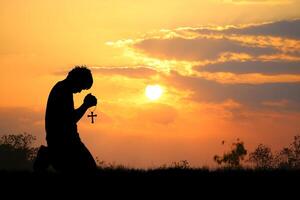 Silhouette of Person Praying with Cross at Sunset in Tranquil Outdoor Setting with Vibrant Sky and Clouds photo