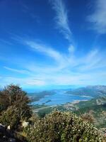 Wide-Angle View of the Bay of Kotor, Montenegro from a Mountain Peak, Featuring Coastal Towns, Lush Greenery, Blue Water, and a Vibrant Sky with Thin Clouds on a Clear Day photo