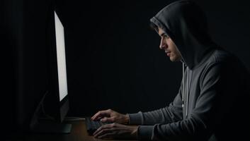 An anonymous figure in a hoodie uses a computer within a dimly lit room evoking a sense of cybercrime intrigue photo