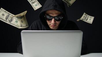 Hooded hacker with sunglasses and laptop amidst cascading dollar bills represents cyber crime online fraud and data theft photo