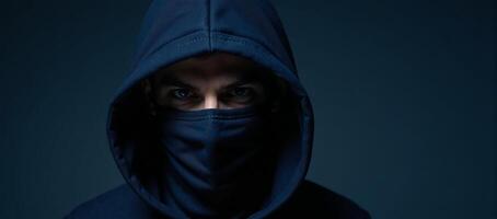A darkhoodied masked man with piercing blue eyes looks straight into the camera photo