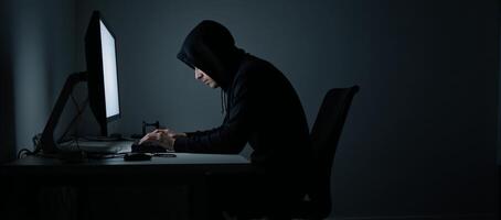 Hacking computer in dim room hooded cybercriminal using malware for device intrusion photo