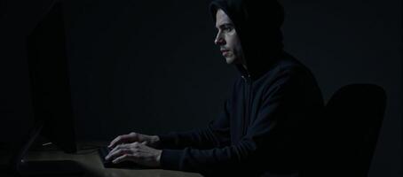 An anonymous figure in a hoodie uses a computer within a dimly lit room evoking a sense of cybercrime intrigue photo