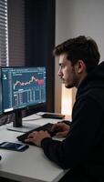 A hooded hacker performs stock analysis at his computer with market data displayed on a nearby phone photo