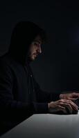 An anonymous figure in a hoodie uses a computer within a dimly lit room evoking a sense of cybercrime intrigue photo