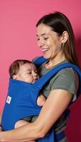 A smiling mother tenderly holds her sleeping baby in a blue carrier on a pink backdrop photo