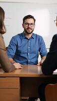 Bearded man discusses with others while seated at a desk with whiteboard and wearing glasses photo