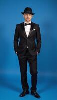 Man in black suit hat stands before blue backdrop photo
