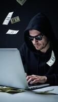 Hooded hacker with sunglasses and laptop amidst cascading dollar bills represents cyber crime online fraud and data theft photo